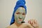 A woman with a brush applies a clay mask to her face with blue towel on her head