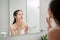 Woman caring of her beautiful skin on the face standing near mirror in the bathroom. Young woman applying moisturizer on her face