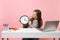 Woman in casual clothes hold looking on round alarm clock while sit work at white desk with contemporary pc laptop