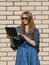 Woman in casual wear leans against white brick wall. Red hair girl is standing outside an office, holding a tablet