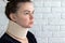 Woman with cervical surgical collar feeling pain in neck in home against a blurred white brick wall background