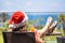 Woman in christmas santa claus hat and slippers relaxing and drinking cocktail on balÑony opposite palm trees beach and sea.