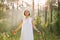 Woman with closed eyes in white nightgown standing in green forest at sunrise