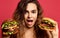 Woman compare big beef burger sandwich and huge cheeseburger with hungry mouth happy shouting on pink red background