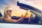 Woman in convertible car taking selfie over sunset
