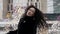 A woman with curly hair in a black fur coat, walks against the background of modern office buildings and against the