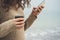 Woman with curly hair in a brown sweater walks on the beach, using cell phone and drinking coffee