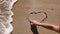 Woman draws heart on a wet sand. Female`s hands writing the word sea on sand. Sea water washes away the drawing on a sandy beach.