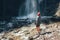 Woman dressed active trekking clothes standing near the mountain river waterfall and enjoying the splashing Nature power.