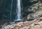 A woman dressed in a red hat and active trekking clothes standing near the mountain river waterfall rose arms up and enjoying the