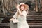 Woman dressed in a white dress holding a book wearing a sunhat on a bridge with a blurry brown colored background