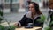 Woman is drinking coffee in street cafe, sitting at table , wearing wireless headphones and turning on music in