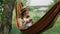 Woman in earphones lying in hammock and listening to music, singing and relaxing at green garden. Cute smiling girl in hat and dre