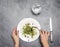 Woman eating fresh green sprouts microgreens and seeds in white plate on grey background with silverware and copy space.  Hands.