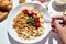 Woman eating healthy breakfast bowl, hold in hand granola, seeds, fresh strawberry, banana, top view, copy space.