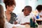 Woman Elementary School Teacher Giving Male Pupil Wearing Uniform One To One Support In Classroom