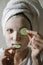 A woman in a face mask is having fun with round cucumbers, looking at the camera and biting a cucumber. Natural