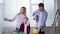 woman with female child watch as man mixes pigment with paint in bucket using construction drill in apartment during