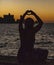 Woman forms heart with her hands, while sitting on a pier at sundown