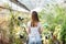 Woman gardener in apron caring potted plant in greenhouse surrounded by plants and pots. Home gardening, love of plants and care.