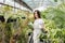 Woman gardener in apron caring potted plant in greenhouse surrounded by plants and pots. Home gardening, love of plants and care.