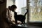 Woman girl in a protective black medical mask, looks at a cat, stands at home near the window during quarantine covid-19