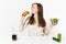 Woman with green detox smoothies, salad in glass bowl, cucumber, greedily eats burger, cola in bottle isolated on white