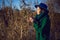 woman in a green knitted sweater and a blue hat stands by the bushes at sunset and drinks tea from an iron thermos mug