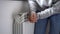 Woman in grey sweatshirt puts hands on room central heating battery to warm up in cold apartment