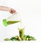 Woman hand with blender pouring green smoothie drink in glass with splashing at white background with green ingredients, fruits