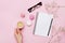 Woman hand hold cup of coffee, cake macaron, clean notebook, eyeglasses and flower on pink table from above. Female working desk.