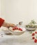 Woman hand holding bowl with vegan porridge and  fresh raspberries above kitchen table with bowls and ingredients at white wall