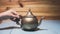 Woman hand holding handle of vintage copper teapot kettle with blemishes and marks, sitting in blue wooden table.
