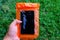 Woman hand holding the orange waterproof mobile phone case over the green grass background. PVC zip lock bag protect mobile phone