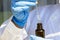 Woman hand in white coat and blue medical gloves holding pipette dropper with drop of natural oil above brown glass bottle