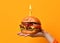 Woman hands hold big burger barbeque sandwich with beef and lit candle for birthday party on yellow