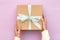 Woman hands holding craft gift box with blue bow on pastel pink background. Girl binds classic bow for present box. Top view, flat