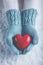 Woman hands in light teal knitted mittens are holding beautiful glossy red heart in snow background