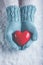Woman hands in light teal knitted mittens are holding beautiful glossy red heart in snow background. Love, St. Valentine concept