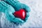 Woman hands in light teal knitted mittens are holding a beautiful glossy red heart in a snow background. St. Valentine concept.