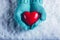 Woman hands in light teal knitted mittens are holding a beautiful glossy red heart in a snow background. St. Valentine concept.