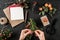 Woman hands making Christmas wreath on dark background with paper card note, pencil, scissors, fir branches and berries.