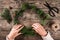 Woman hands making a Christmas wreath of fir branches and pine cones on wooden background with scissors and skein of jute