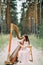 Woman harpist sits at forest and plays harp against a background of pines