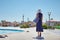 Woman in hat and blue dress go for a walk in the hotel resort near swimming pool with beach bag