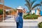 Woman in hat and blue dress go for a walk in the hotel resort near swimming pool with beach bag