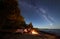 Woman having a rest at night camping near tourist tent, campfire on sea shore under starry sky