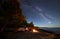 Woman having a rest at night camping near tourist tent, campfire on sea shore under starry sky