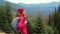 Woman hiker with backpack, wearing in red jacket and orange pants, standing on the mountains and forest background