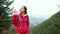 Woman hiker with backpack, wearing in red jacket and orange pants, standing on the mountains and forest background